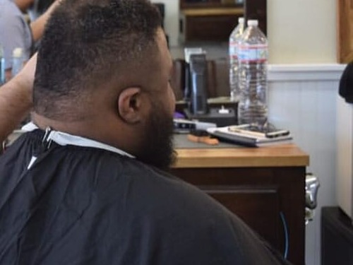 Back view of a bearded man in a barber cape seated at the shop station